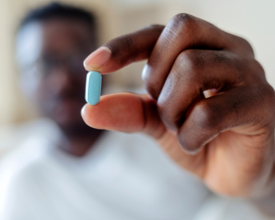 Black man blurred in background holding his hand out which is in focus holding a blue pill